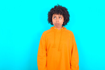 Fototapeta na wymiar Dissatisfied young woman with afro hairstyle wearing orange hoodie against blue background purses lips and has unhappy expression looks away stands offended. Depressed frustrated model.