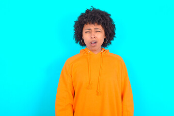 Obraz na płótnie Canvas young woman with afro hairstyle wearing orange hoodie against blue background yawns with opened mouth stands. Daily morning routine