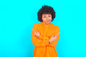 Obraz na płótnie Canvas young woman with afro hairstyle wearing orange hoodie against blue background crosses arms and points at different sides hesitates between two items or variants. Needs help with decision