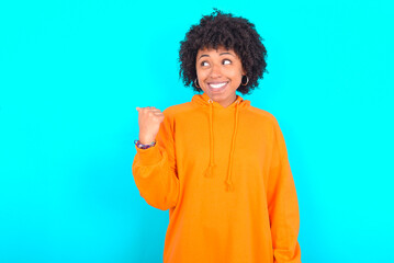 Obraz na płótnie Canvas young woman with afro hairstyle wearing orange hoodie against blue background points away and gives advice demonstrates advertisement