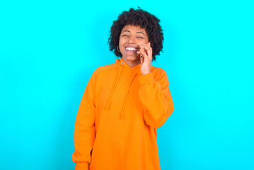Fototapeta na wymiar Funny young woman with afro hairstyle wearing orange hoodie against blue background laughs happily, has phone conversation, being amused by friend, closes eyes.