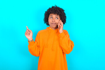 young woman with afro hairstyle wearing orange hoodie against blue background speaks on mobile phone spends free time indoors calls to friend.