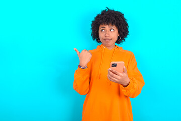 Obraz na płótnie Canvas young woman with afro hairstyle wearing orange hoodie against blue background points thumb away and shows blank space aside, holds mobile phone for sending text messages.