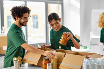 charity, donation and volunteering concept - happy smiling volunteers packing food in boxes at...