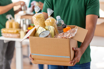 charity, donation and volunteering concept - close up of male volunteer's hands holding box with food over group of people at distribution or refugee assistance center - obrazy, fototapety, plakaty
