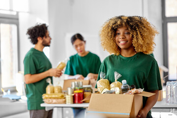 charity, donation and volunteering concept - happy smiling female volunteer with food in box and international group of people at distribution or refugee assistance center