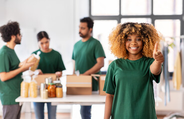 charity, donation and volunteering concept - happy smiling female volunteer pointing to camera and international group of people packing food in boxes at distribution or refugee assistance center