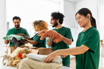 charity, donation and volunteering concept - international group of happy smiling volunteers packing clothes and other stuff in boxes at distribution or refugee assistance center