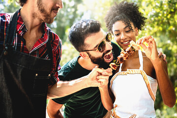 Multiethnic Joyful young people joking outdoors at barbecue eating meat skewers and having fun...