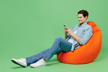 Full size happy young brunet man 20s years old wear blue shirt sit in bag chair hold in hand use...
