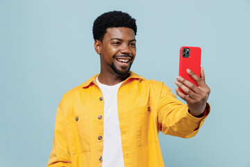 Young happy man of African American ethnicity 20s wear yellow shirt doing selfie shot on mobile cell phone post photo on social network isolated on plain pastel light blue background studio portrait