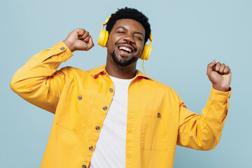 Young cheerful fun cool man of African American ethnicity 20s in yellow shirt headphones listen to music dance isolated on plain pastel light blue background studio portrait. People lifestyle concept.