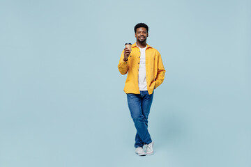 Full body fun young man of African American ethnicity 20s wear yellow shirt hold takeaway delivery craft paper brown cup coffee to go isolated on plain pastel light blue background studio portrait