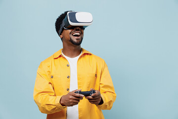 Young man of African American ethnicity 20s wear yellow shirt hold in hand play pc game with joystick console watching in vr headset pc gadget isolated on plain pastel light blue background studio