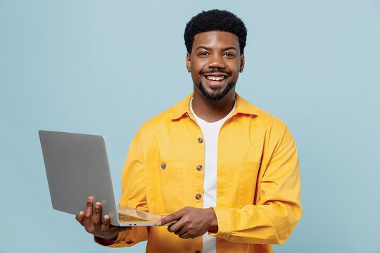 Young cheerful happy man of African American ethnicity 20s in yellow shirt hold use work on laptop pc computer isolated on plain pastel light blue background studio portrait. People lifestyle concept.