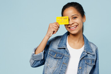 Little happy kid teen girl of African American ethnicity 12-13 years old in denim jacket hold in hand cover eye with credit bank card isolated on pastel plain light blue background Childhood concept.