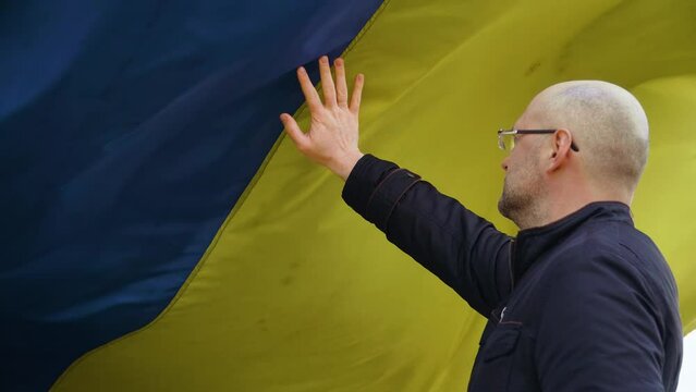 Ukrainian man patriot touches large developing flag of his country during armored invasion. Crisis, peace, stop aggression in whole world