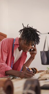 Vertical video of happy african american leather craftsman with dreadlocks on the phone at workshop