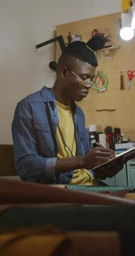 Vertical video of african american leather craftsman with dreadlocks writing notes in workshop