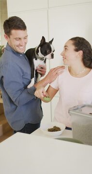 Vertical video of smiling caucasian couple holding pet dog and measuring out dog food into a bowl