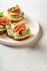 Bruschetta with baguette, bacon or meat, cream cheese, micro-greenery, fresh cucumber and sprouts, on white plate on white textured background. Text space