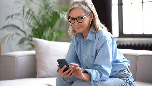Smiling senior lady using smartphone texting message sit on couch, middle aged woman grandma hold phone typing sms enjoying communication in mobile app at home, older people and modern tech concept