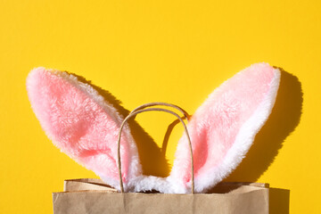 Bunny ears stick out of a paper bag on a yellow background. Holiday shopping concept. Flat lay, place for text.