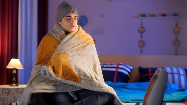 An attractive Indian boy covering himself with a blanket while sitting in front of a room heater - cold climate  a cozy room. A young man wearing a woolen cap  jacket  and a pair of gloves - freezi...