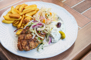 Bifteki, greek minced meat steak stuffed with feta cheese, served with french fries, coleslaw, tzatziki and onions on a white plate, wooden outdoor table, selected focus