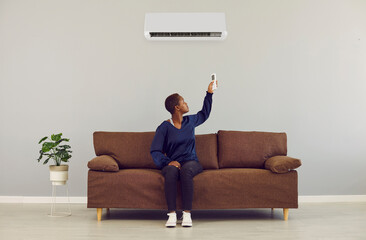 Happy black woman sits on brown couch relaxing under air conditioner at home holding remote control...