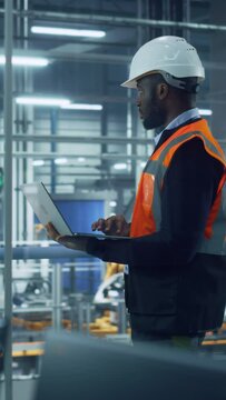 Vertical Portrait of a African American Engineer Using Laptop in Office at a Car Assembly Plant. Vehicle Factory Line Operator Working on Computer, Overviewing Autonomous Electric Car Production.