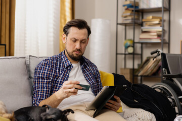 A man in a plaid shirt is relaxing on a couch with small dog. The man holds a tablet in his hand...