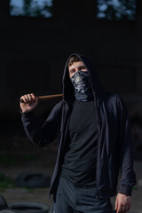 Naklejka na ściany i meble A young tough guy in a buff mask and hood threatens the viewer with a rusty iron bat or a baton with a chain. Hooligan, robber, aggressive anarchist, bully or resistance fighter, system protester