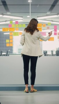 Vertical Portrait of a Young Confident Businesswoman Creating Project Plan on Office Wall with Paper Notes. Manager Working on Business, Financial and Marketing Projects. Specialist in Diverse Team.