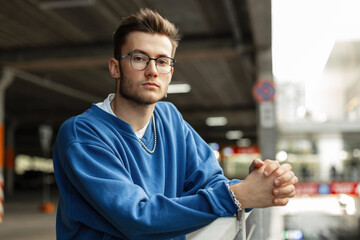 Fashion handsome man with hairstyle and stylish glasses in blue sweatshirt stands and looks at the...