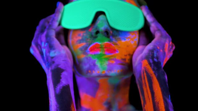 Beautiful young woman in neon light. Portrait of a model with fluorescent makeup posing in ultraviolet light with colorful makeup, wearing glasses and dancing.