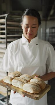 Vertical video of portrait of happy asian female baker with bread in bakery