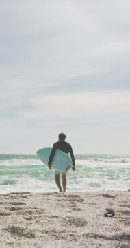 Vertical video of biracial man alone with surfboard on sunny beach