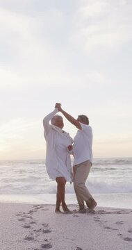 Vertical video of happy senior biracial couple dancing on sunny beach