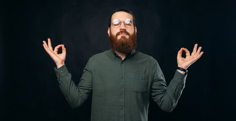 Young bearded man is meditating while holding his hand up with zen gesture. Studio shot over black background.