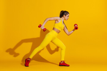 Studio shot of sportive slim girl workout with sports equipment isolated on bright yellow studio background with shadow. Beauty, sport, action, fitness, youth concept.