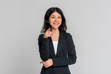 Happy young dark-haired girl in black blazer smiling and looking camera while pointing to you,...