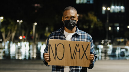 Portrait angry african american man in face mask stands in city at night evening holding sign...