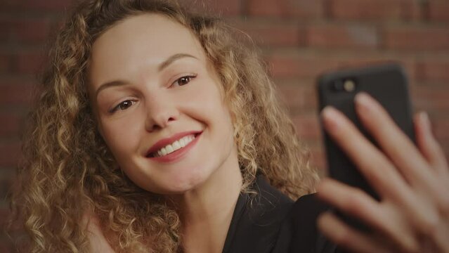 Portrait of a young curly caucasian business woman taking selfies and smiling with a red brick wall