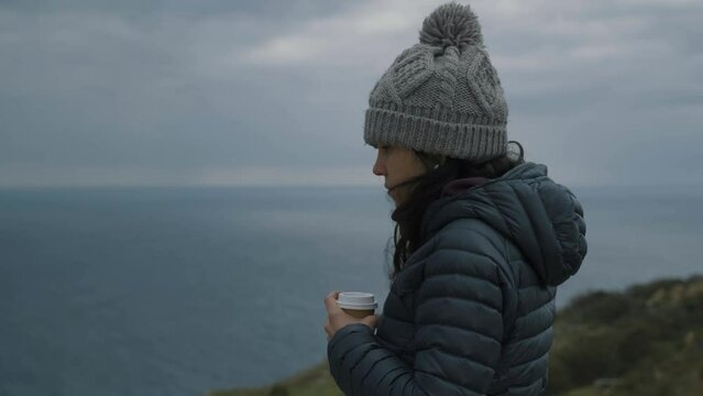 Young woman enjoying coffee and reflecting and contemplating by the sea shore on a cold and cloudy winter day, Malta