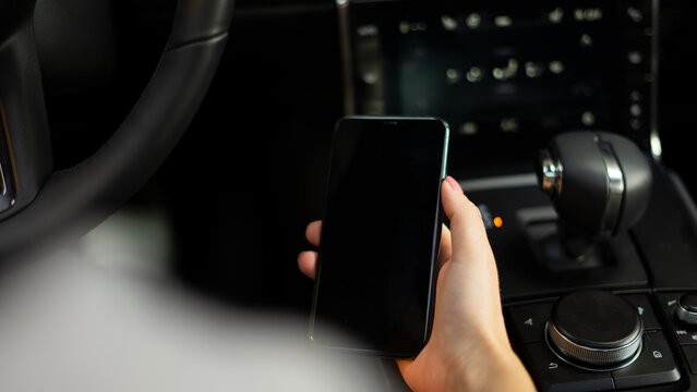 Eine Person h&auml;lt ein Mobiltelefon in der Hand, im Hintergrund ist ein Lenkrad und ein Multiinformationsdisplay eines Fahrzeuges zu sehen.