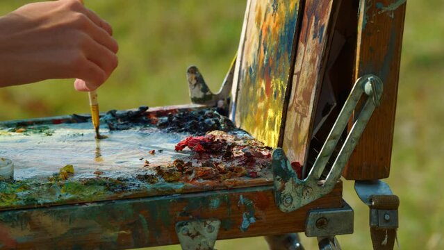 Painter hands drawing picture on easel closeup. Artist mixing paints on palette.