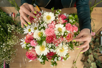Top view of the hands of a young female florist creating a beautiful composition of delicate pink roses, carnations and white daisies on the table