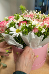 Close-up of the hands of a young female florist packing a beautiful composition of delicate pink roses, carnations and white daisies into a cardboard box and paper on the table. Vertical photo