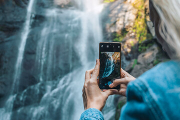 Close-up of a young female traveler photographing a waterfall with a mobile phone. First-person...